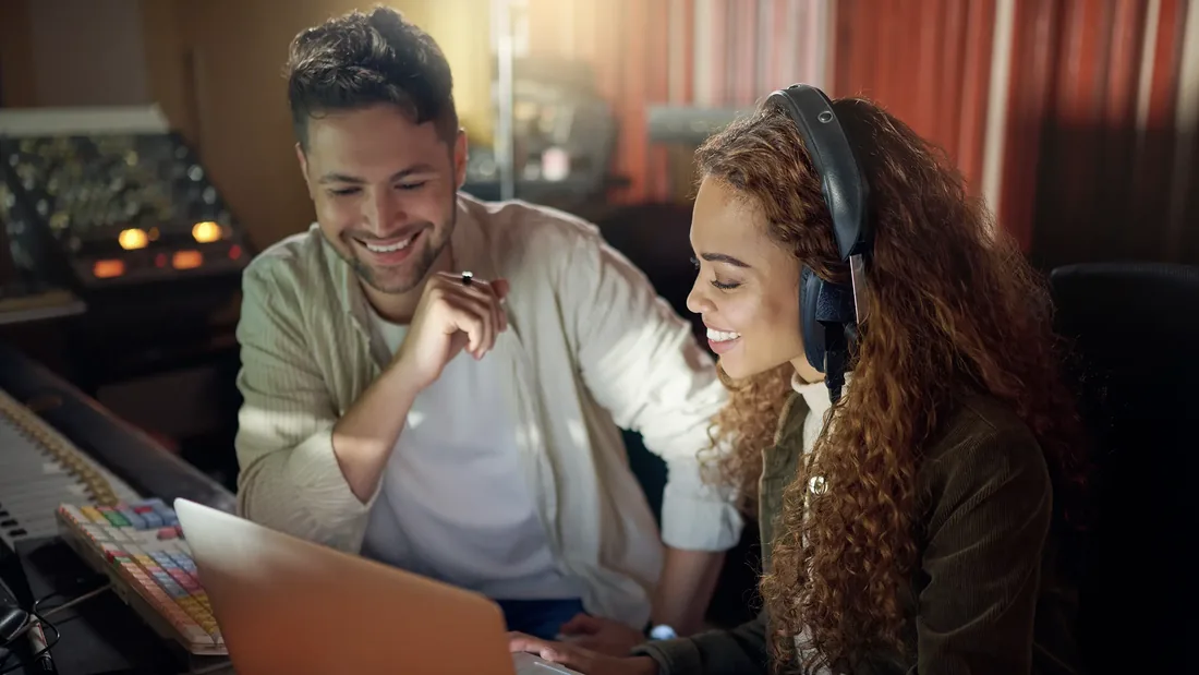 Two musicians in a home studio, smiling at a laptop screen, a keyboard and mixing desk beside them.