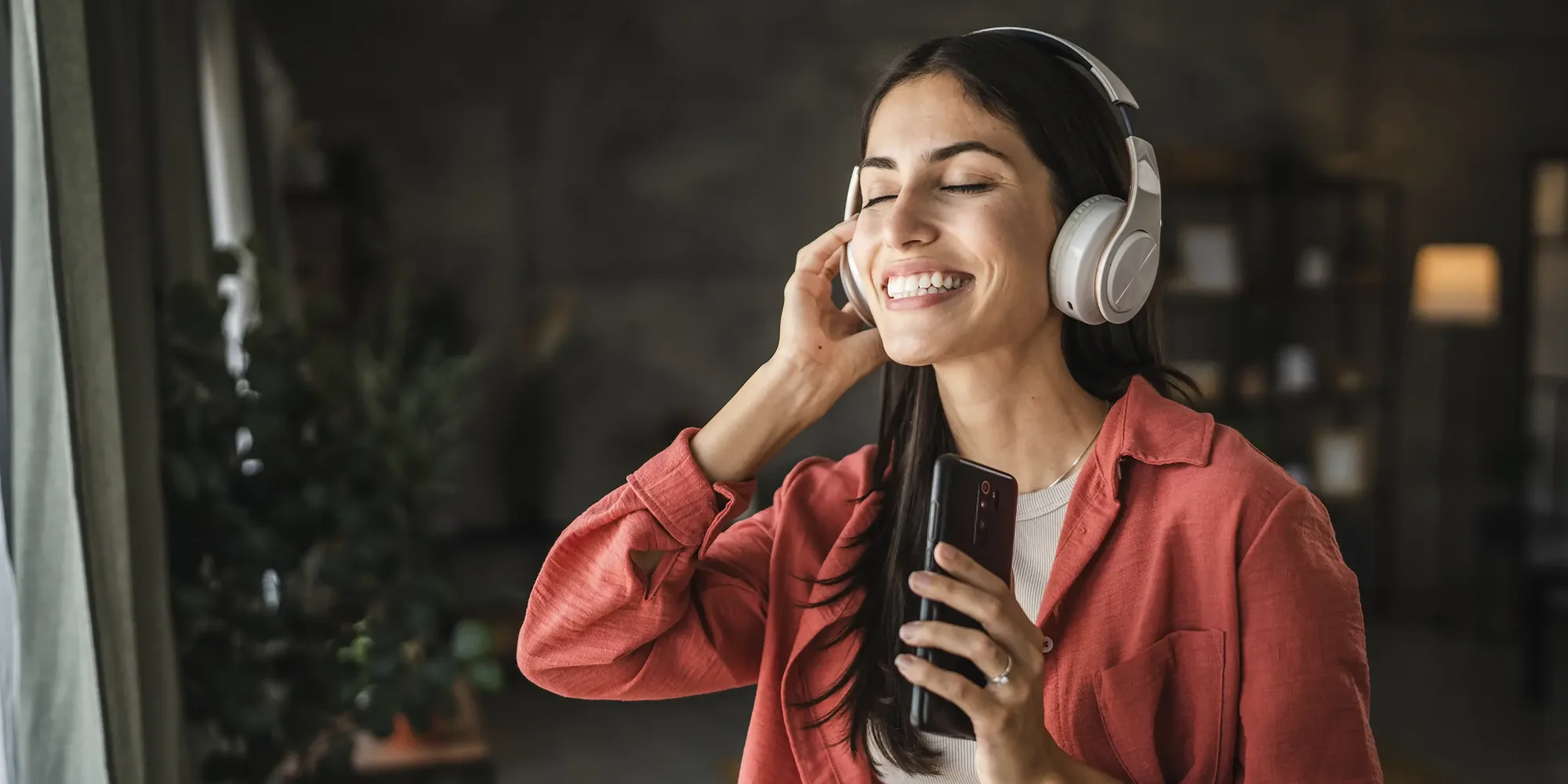 A woman listening on over-ear headphones in a warm, dimly lit room, eyes closed and smiling.